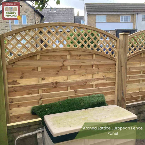 A KDM Arched Lattice Top Fence Panel (ALT180) with durable, pressure-treated timber boards is installed in a backyard, with a green mat and beige utility box placed in front of the fence.