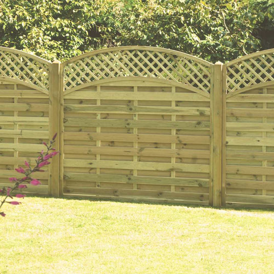 KDMs Arched Lattice Top Panels (ALT180) elegantly stand in a grassy yard with lush trees as a backdrop and pink-flowered branches enhancing the charm. Constructed from pressure-treated wood, these panels promise durability and beauty across seasons.