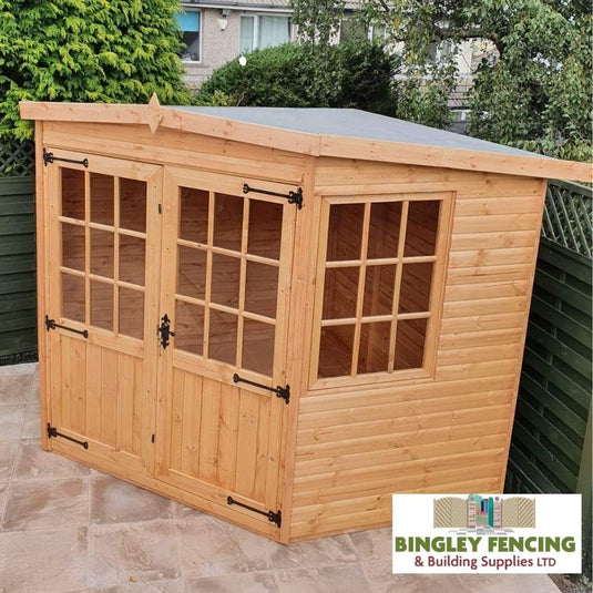 This elegant wooden garden shed, featuring double glass-paneled doors and a side window, enhances any patio with its slanted roof. Surrounded by green fencing and trees, it becomes a cozy Corner House - Summerhouse by Bingley Fencing. The corner is adorned with the Bingley Fencing & Building Supplies LTD logo.