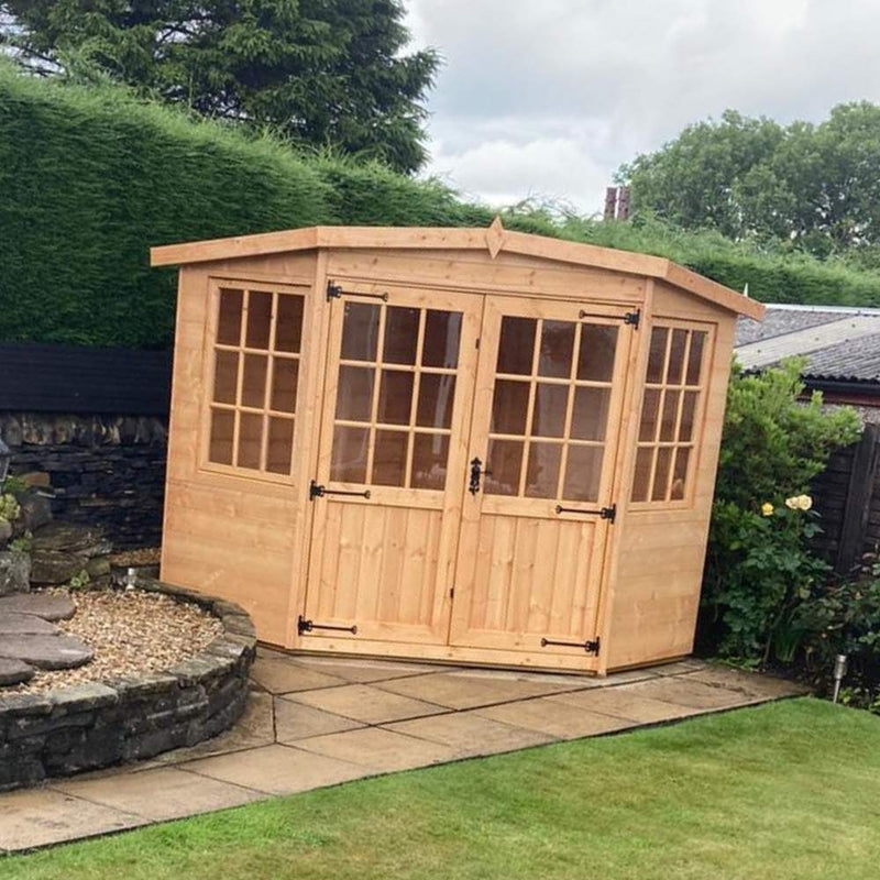 Load image into Gallery viewer, Wooden garden sheds sit on a paved area surrounded by greenery. The custom-designed Bingley Fencing Corner House - Summerhouse showcases double doors with glass panels and windows on either side, nestled near a stone wall and lush bushes under a partly cloudy sky.