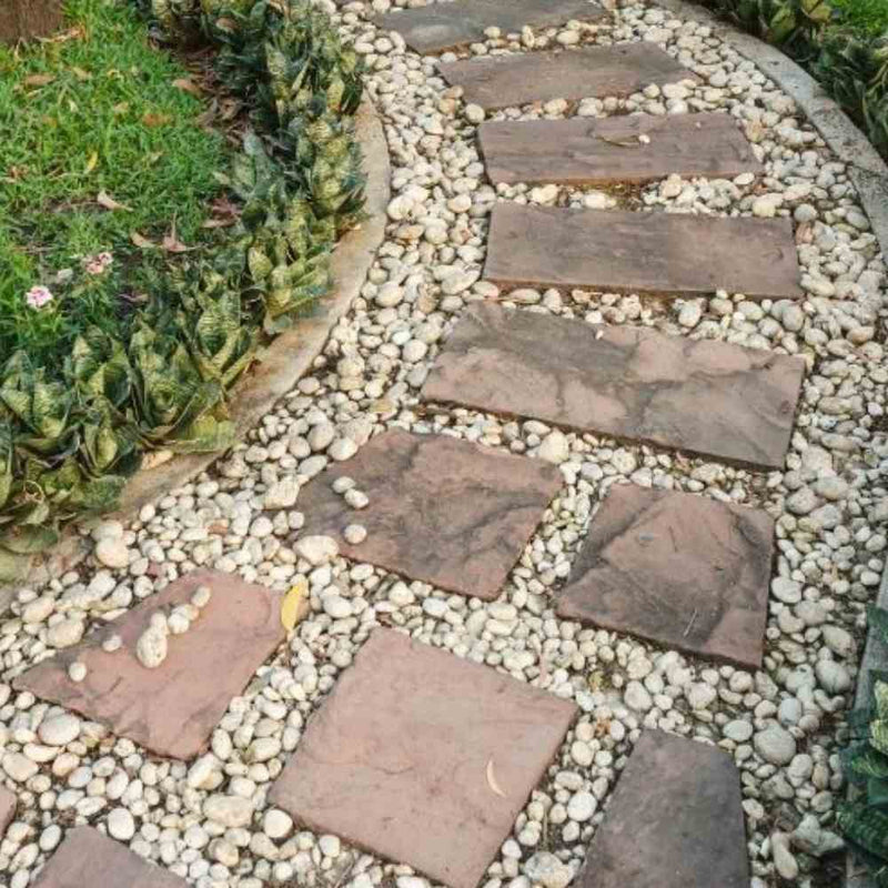 Load image into Gallery viewer, A curved garden pathway composed of large, rectangular stone slabs is elegantly set in a bed of 40mm pieces from The Heritage Stone Company's Blue Slate - 20kg. On the left side, a border of green plants with jagged leaves runs alongside a grassy area, enhancing the decorative garden ambiance.