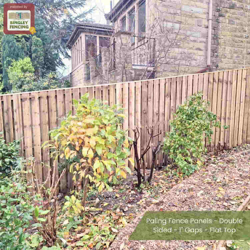 A Bingley Fencing wooden paling fence with double-sided, flat top panels and 1 gaps lines a sloping garden with shrubs. A stone house and trees are visible behind. Text: Paling Fence Panels - Double Sided - 1 Gaps - Flat Top.