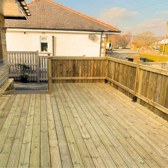 Wooden deck with railings attached to a house, overlooking a fenced yard. There is a small potted plant near the door and another house visible in the background under a partly cloudy sky.