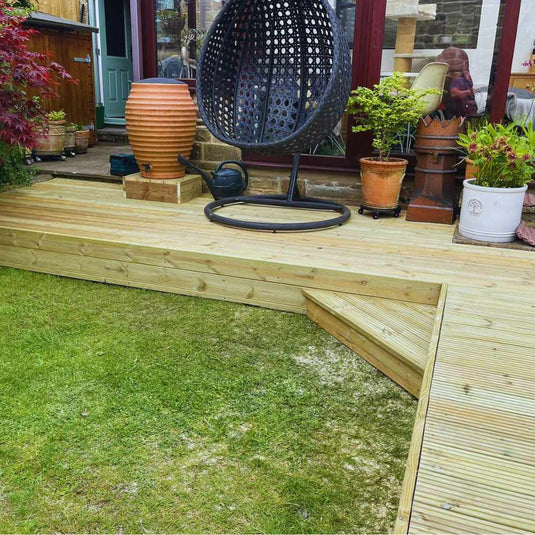 A wooden deck with a small step leads to a grassy lawn. On the deck are potted plants, a large terracotta jar, and a black hanging egg chair near glass doors of a house.