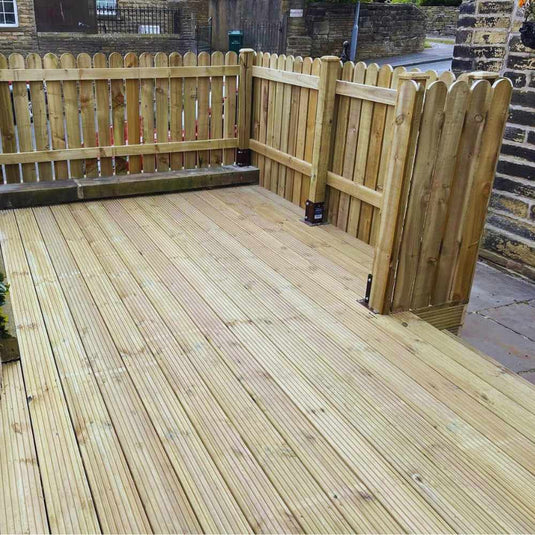 A wooden deck with a light brown finish is enclosed by a wooden picket fence, situated outdoors near a stone wall and a street in the background.