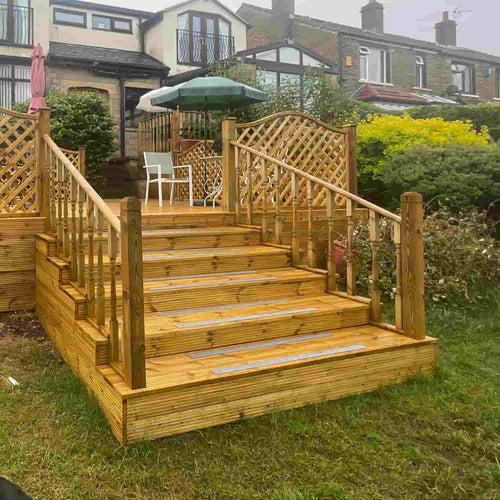 A wooden deck with KDM Decking Handrail & Fillet - 12' Lengths, smooth timber steps, and lattice panels leads to a garden seating area with a white chair and green umbrella, behind a house bordered by bushes, grass, and neighboring homes.