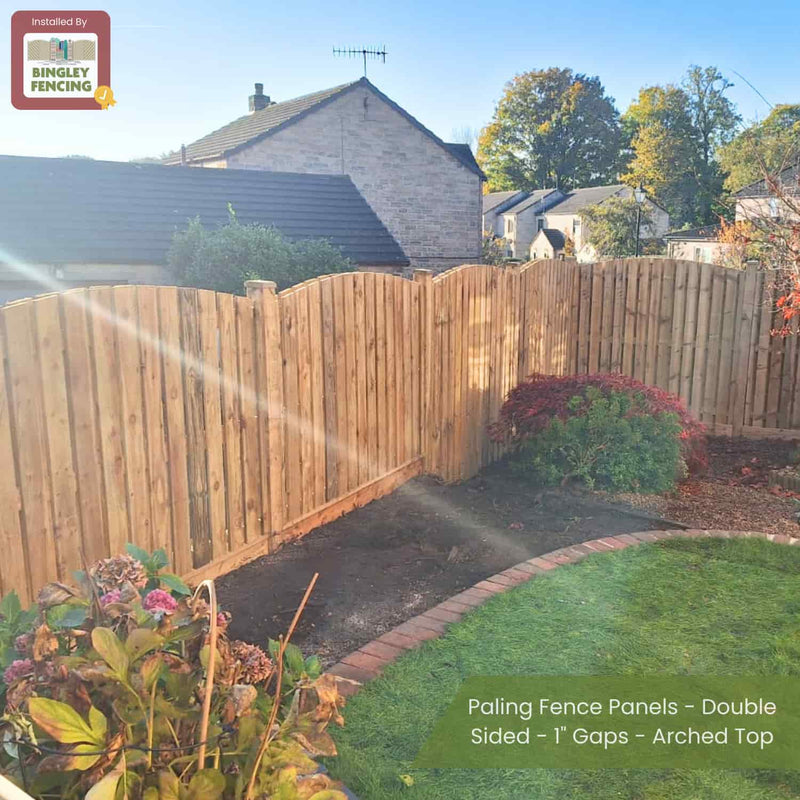Load image into Gallery viewer, A garden with Bingley Fencing Paling Fence Panels—Double Sided, 1 Gaps, Arched Top—bordered by short red brick edging and flowers; houses and trees are visible in the sunny background.