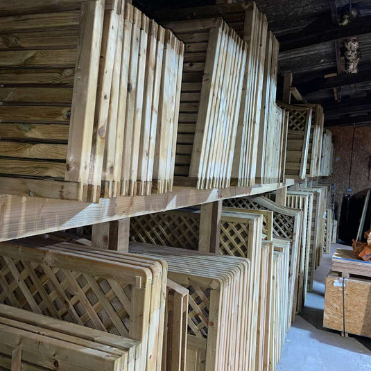 Stacks of wooden fence panels and lattice sections are organized on shelves in a storage area. The wood grain and texture are visible, and the panels are neatly arranged, showcasing their geometric patterns.