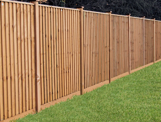Alongside a neatly manicured green lawn stands a long Bingley Fencing Featheredge Fence Panel - Flat Top, crafted with vertical wooden panels. The Tanalised finish ensures durability, while dense, dark foliage in the background offers contrast and privacy to the lighter colors of the fence and grass.