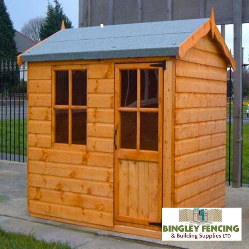 Load image into Gallery viewer, The Hobbit Kids Playhouse, by Bingley Fencing, is a small wooden structure featuring a window and door, topped with a slanted gray roof. It is placed on a concrete surface and treated with child-friendly preservatives. A metal fence and surrounding greenery provide the setting, with the Bingley Fencing logo displayed at the bottom.