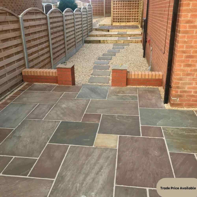 Load image into Gallery viewer, A backyard features a path composed of Bingley Fencing's Indian Stone Paving - Project pack, which includes multicolored Raj Green stone tiles, leading to a raised area bordered by brick planters. Fences line both sides of the path.