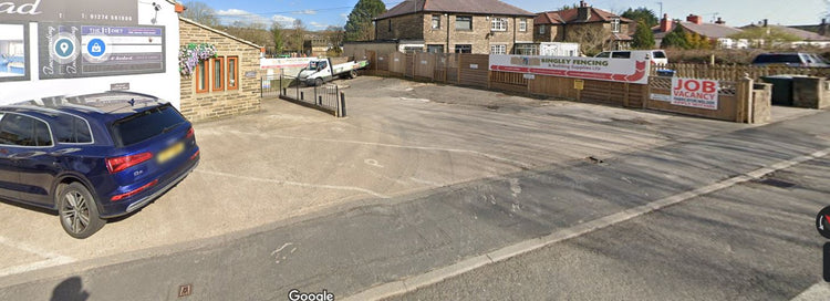 A small parking area outside a business with signs for Fencing and Job Vacancy. A few cars are parked, and there are stone buildings and a wooden fence in the background under a blue sky.