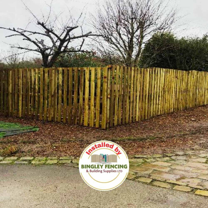 Load image into Gallery viewer, A garden area is enclosed by Paling Fence Panels - Single Sided with 2" gaps and a flat top, surrounded by bare trees and bushes on a cloudy day. The ground is scattered with fallen leaves and features a cobbled path. In the foreground, the brand name Bingley Fencing is prominently displayed, indicating their installation services.