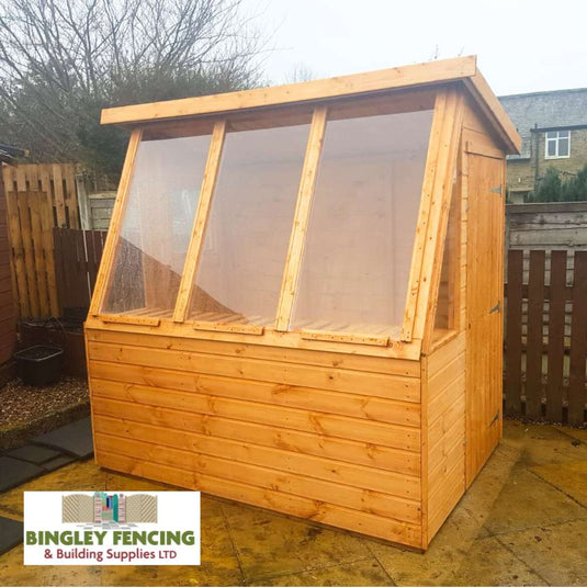A beautifully displayed custom-made structure, "The Potting Shed - Gardeners Choice," features a slanted roof and large clear windows. Positioned on a paved area and surrounded by a wooden fence, this shed is ideal for creating growing space. It proudly displays the Bingley Fencing logo in the bottom left corner.