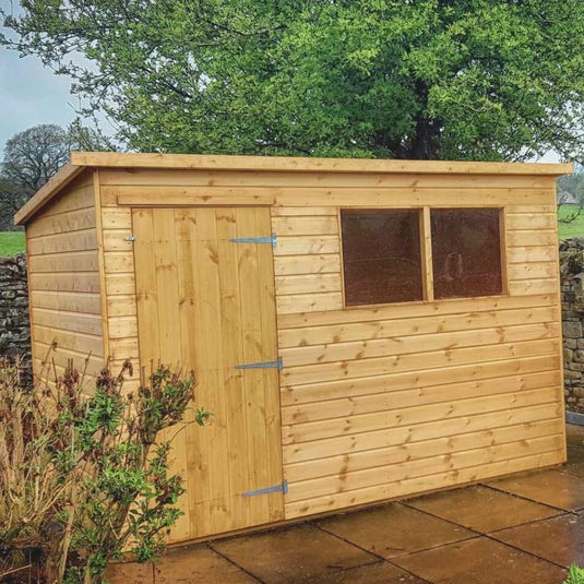 A wooden garden shed with a slanted roof, single door, and two rectangular windows stands on a paved patio with greenery and a tree in the background.