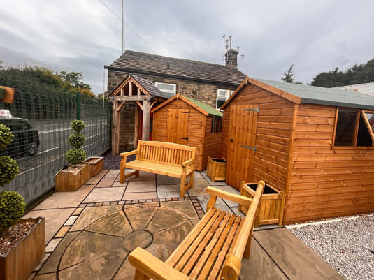A small fenced garden with wooden sheds, benches, potted plants, a stone patio, and a stone building in the background under a cloudy sky.