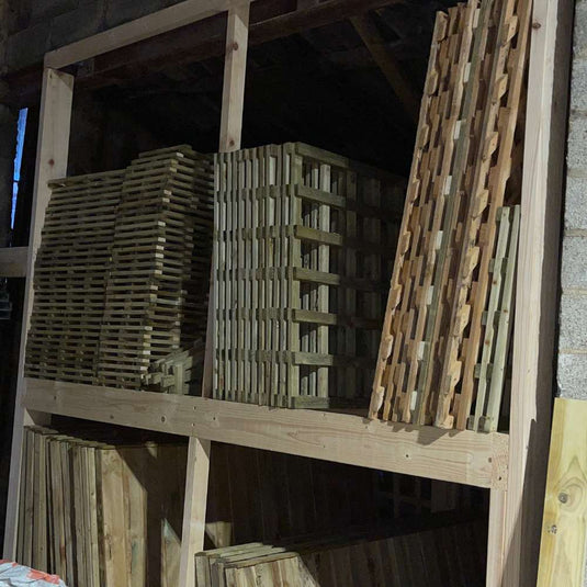 Stacks of wooden slats and panels are neatly arranged on a wooden shelf in a dimly lit storage area with brick walls.