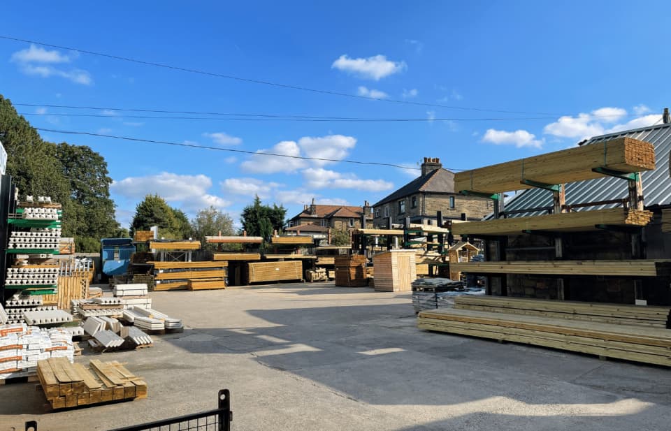 Outdoor lumberyard under a blue sky with scattered clouds. Several stacks of timber and building materials are organized on racks. A few residential houses are visible in the background, surrounded by trees.