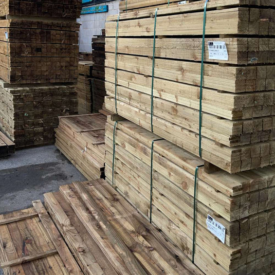 Stacks of wooden planks and boards are piled and bundled together with green straps in an outdoor lumber yard. Some pieces have labels attached. The wood appears to be treated and is neatly organized.