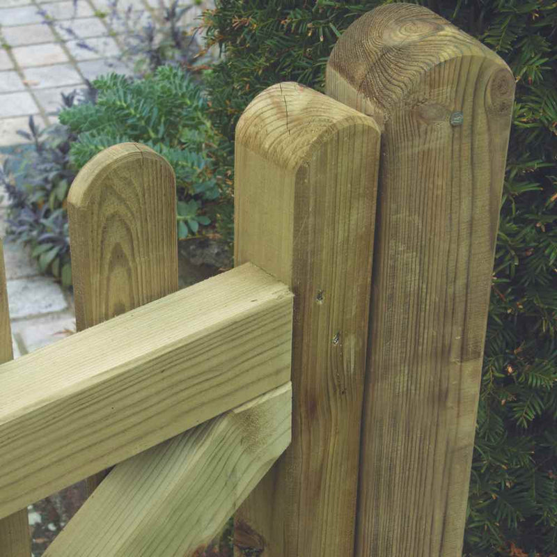 Load image into Gallery viewer, The KDM Premium Round Top Picket Gate - PPG90 is shown in a close-up, featuring rounded posts and a decorative diagonal brace. Pressure-treated with a natural finish to enhance its rustic look, it blends seamlessly into the landscape, surrounded by greenery leading to a paved area.