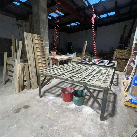 A workshop with a large metal-framed workbench. Wooden pallets are stacked against a wall. Red and green buckets are on the floor. Overhead, red coiled cables hang from the ceiling. The space is dimly lit and cluttered.