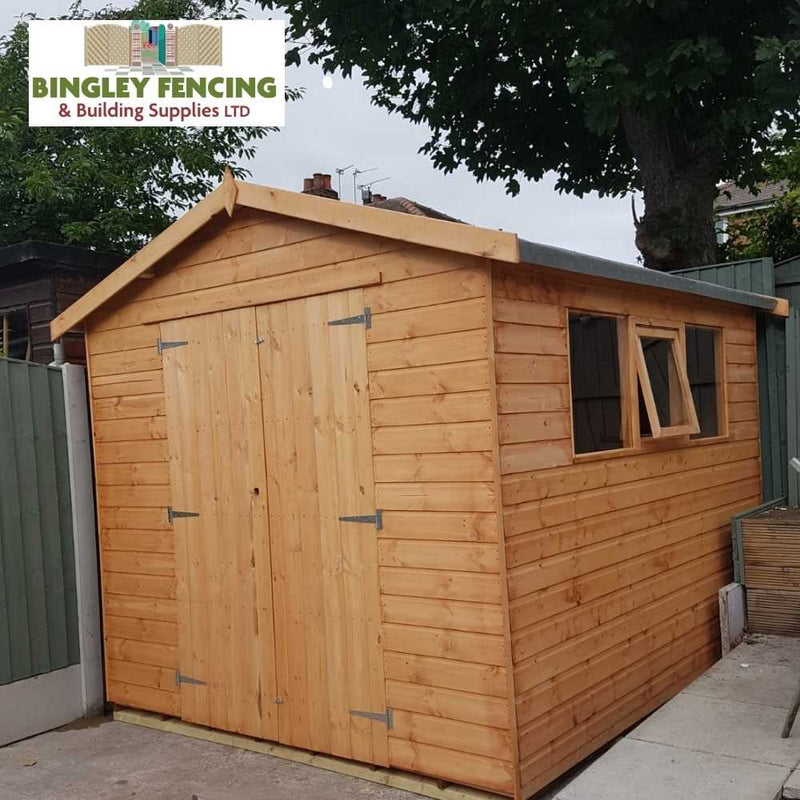 Load image into Gallery viewer, A personalized "The Workshop - Speciality Shed" by Bingley Fencing, featuring a wooden structure, pitched roof, and two windows, is situated on a paved area enclosed by a green fence. Bingley Fencing's logo in the top-left corner guarantees both charm and secure storage for your essentials.