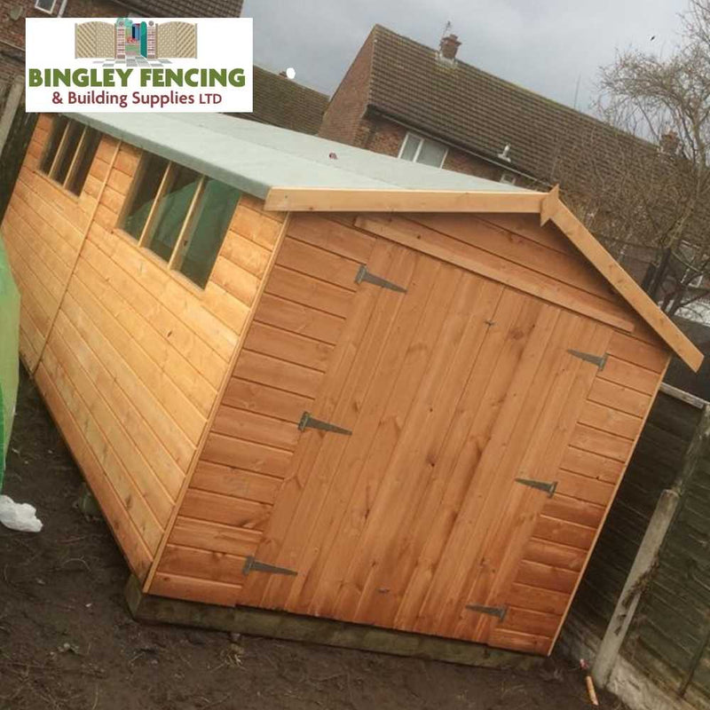 Load image into Gallery viewer, The Workshop - Speciality Shed, crafted from wood and featuring a slanted roof with metal hinges on the door, provides secure storage. It is positioned on muddy ground beside a wooden fence, with a brick house and cloudy sky as the backdrop. In the top left corner is the logo: Bingley Fencing.