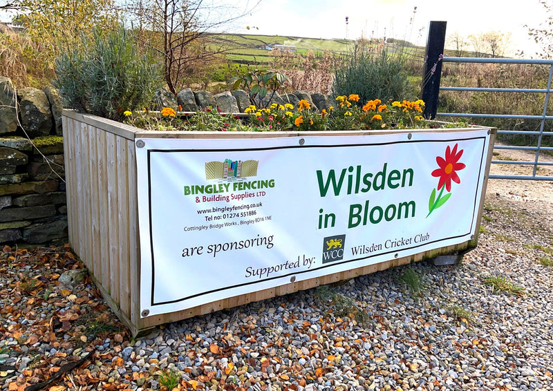 Load image into Gallery viewer, A bespoke tall planter from KDM, crafted using moisture-resistant tanalised timber, is brimming with vibrant orange marigold flowers. It prominently features a "Wilsden in Bloom" sign, sponsored by Bingley Fencing and Wilsden Cricket Club. In the background stretches a picturesque rural landscape with a stone wall and rolling fields.