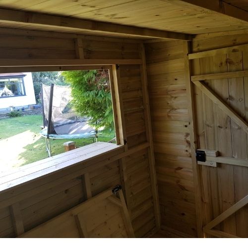 Load image into Gallery viewer, Inside a wooden shed with an open window, the Garden Bar by Bingley Fencing is visible. Outside, a trampoline, tree, and part of a white building can be seen. The shed's door is closed for security, while sunlight brightens the wood interior.
