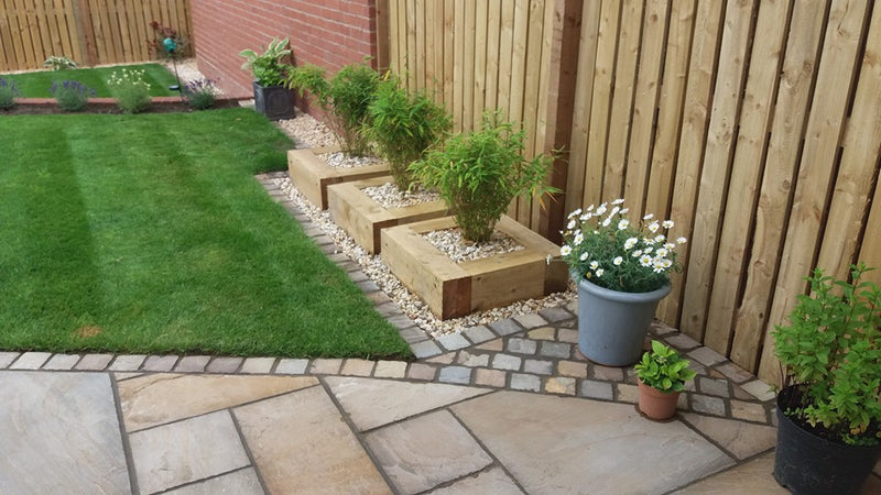 Load image into Gallery viewer, A neatly landscaped backyard showcases a lush green lawn, three wooden planters filled with vibrant plants, and a gravel border. White flowers in pots and a small plant in terracotta containers adorn a stone patio elegantly paved with Bingley Fencing's Indian Stone Paving - Project pack, nestled beside a charming wooden fence.