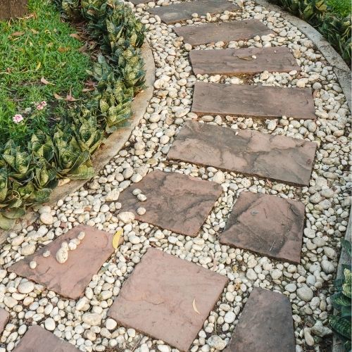 Load image into Gallery viewer, A garden path made of rectangular slabs surrounded by small white pebbles meanders through lush greenery. Green plants with variegated leaves line the path, highlighted by decorative 40mm pieces of Plum Slate from The Heritage Stone Company as ground cover, with grass emerging gently in the background.