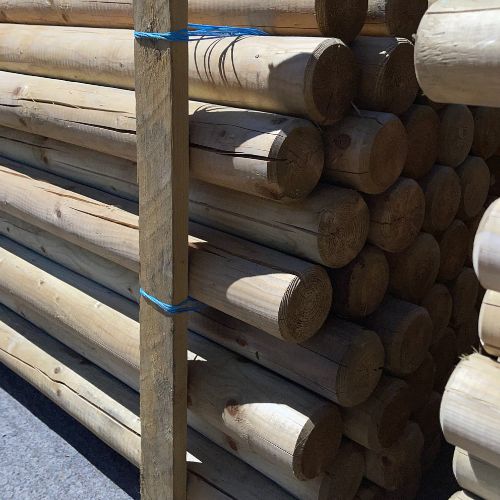 Load image into Gallery viewer, A stack of Bingley Fencing's Tree Stake - Machine Round Spiked Fence Posts, measuring 3” x 8ft, tied together with blue rope, rests on a paved surface. The posts are aligned horizontally, with sunlight casting shadows that highlight their smooth texture and natural grain.