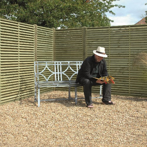 Load image into Gallery viewer, A person wearing a hat sits on a metal bench reading a book in a gravel area, surrounded by KDM Venetian European Fence Panels **Reduced** - VP180, with a tree visible in the background.