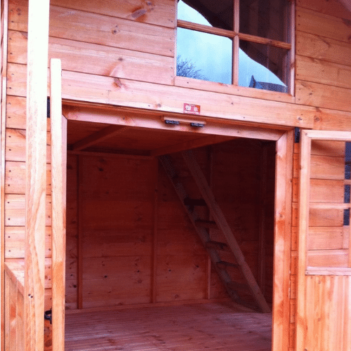 Load image into Gallery viewer, The Adventure Kids Playhouse by Bingley Fencing features open double doors that reveal its interior, complete with a wooden ladder leading to a loft space. Constructed with child-friendly preservatives for safety, the playhouse includes a large window above the entrance to welcome natural light. Its wood features warm tones and visible grain patterns.