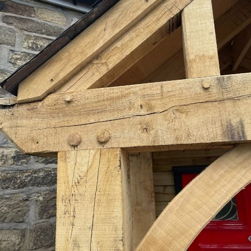 Load image into Gallery viewer, A detailed view of the Entrance Porch in Oak by Bingley Fencing highlights its rustic wooden beams and expertly handcrafted joinery, with wooden pegs securely holding the structure together. In the background, a stone wall is partially visible along with the top of a red door.