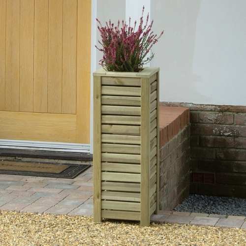 Load image into Gallery viewer, The Reduced Tall Slatted Square Planter by KDM, a tall rectangular planter with horizontal slats, is shown outdoors by a brick wall and wooden door holding a small plant with pink flowers.