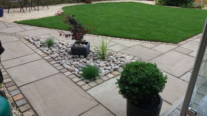 Load image into Gallery viewer, A neatly landscaped garden features a well-manicured lawn surrounded by a stone patio. Decorative stones and potted plants in the center add visual interest, with the elegance enhanced by Bingley Fencing's Indian Stone Paving - Project Pack in Kandla Grey. In the foreground, a small bush sits charmingly in a pot.
