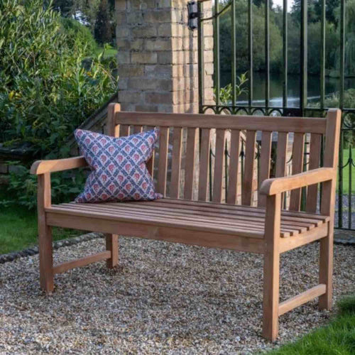 The Bingley Fencing 1.3m Teak Garden Bench, styled in classic West Yorkshire tradition, features a patterned cushion and sits on garden gravel near a brick archway and metal gate, surrounded by greenery and a lakeside view.