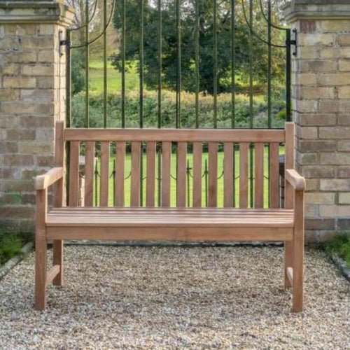 The 1.3m Teak Garden Bench by Bingley Fencing sits on a gravel path between two brick pillars with a black iron gate, set against a backdrop of lush green garden scenery.