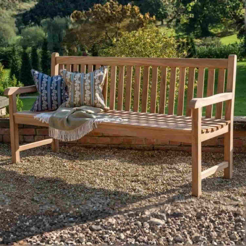 The Bingley Fencing 1.8m Teak Garden Bench sits on gravel in a sunny outdoor space with green grass and trees behind, styled with two patterned cushions and a fringed throw for comfortable, attractive outdoor seating.