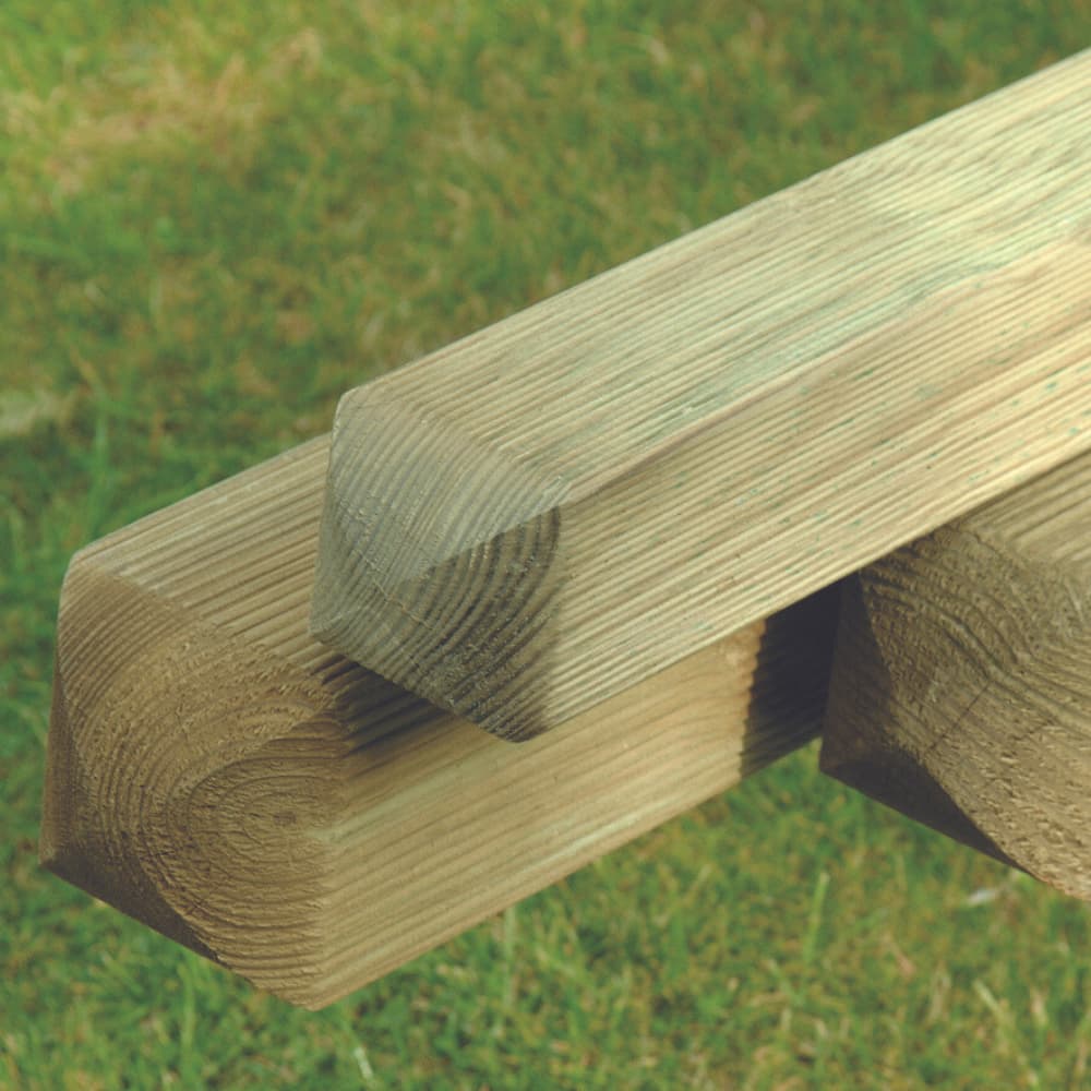 Three wooden fence posts with chamfered (beveled) edges are stacked on each other, resting on a grassy surface. The wood appears to be pressure-treated, showing visible grain lines.