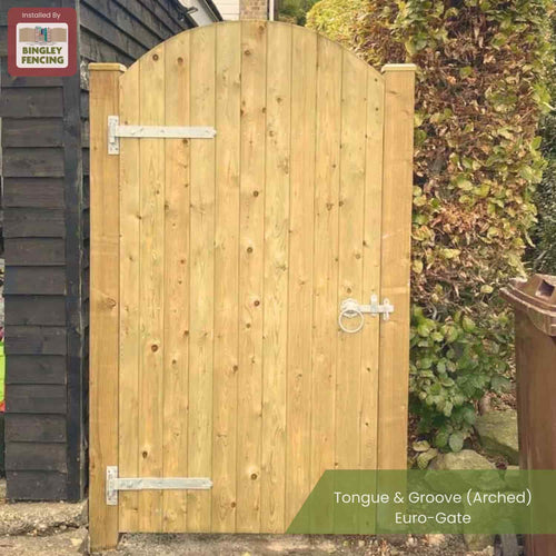 A wooden arched gate with vertical tongue and groove panels and metal hinges, installed between a black shed and leafy bushes; a sign reads Tongue & Groove (Arched) Euro-Gate by Bingley Fencing.