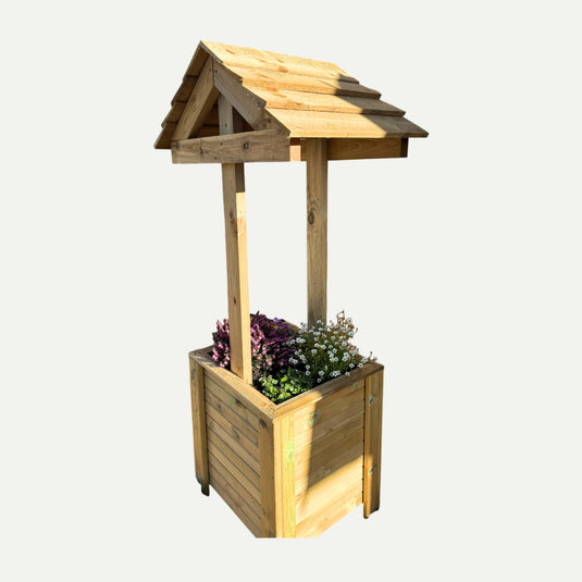 The KDM Wishing Well Planter, crafted from pressure-treated timber with a pitched roof, is filled with green, purple, and white flowering plants and set against a plain white background.