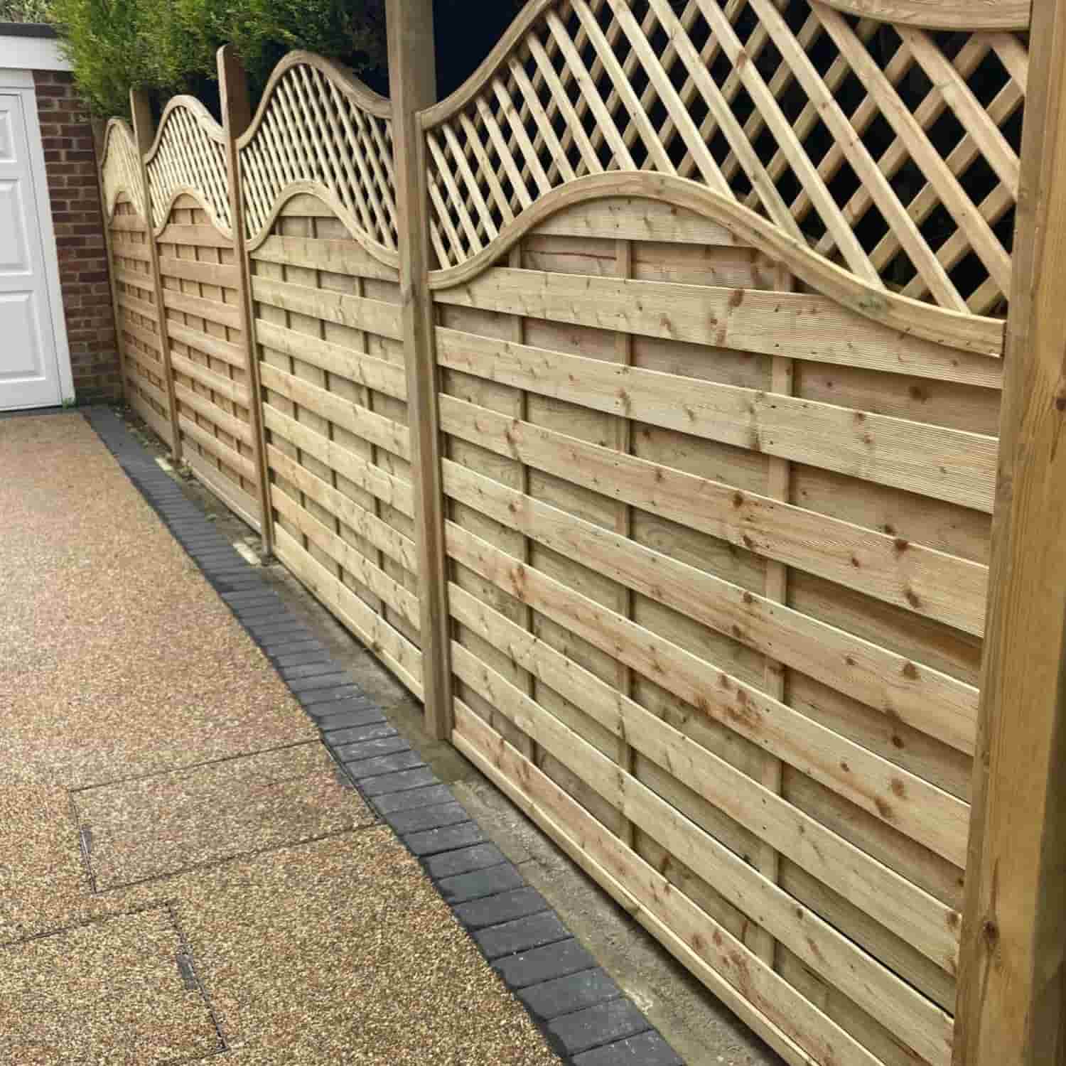 A wooden garden fence with a wavy top edge and lattice design on the upper section, running alongside a paved pathway next to a house.