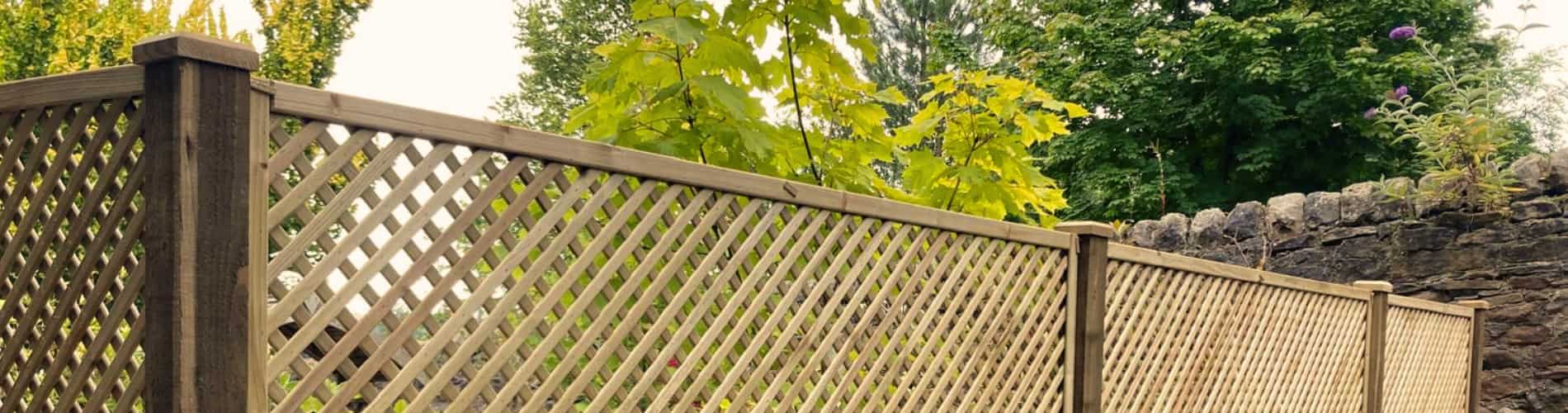 A wooden lattice fence with square-patterned panels stands in a garden, bordered by green trees and shrubs, with a stone wall visible in the background.
