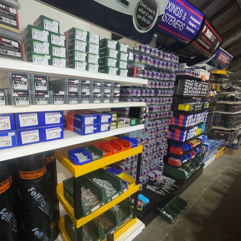 Shelves in a hardware store display various boxes and containers of nails, screws, fasteners, and other building supplies, organized by type and size, with colorful labels and signs above each section.