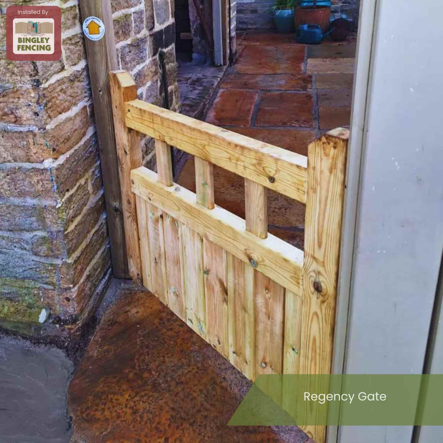A KDM Regency Euro-Gate, made of pressure-treated timber with vertical and horizontal slats, is fitted between a stone wall and metal post on a stone path. A Bingley Fencing sign is displayed on the wall.