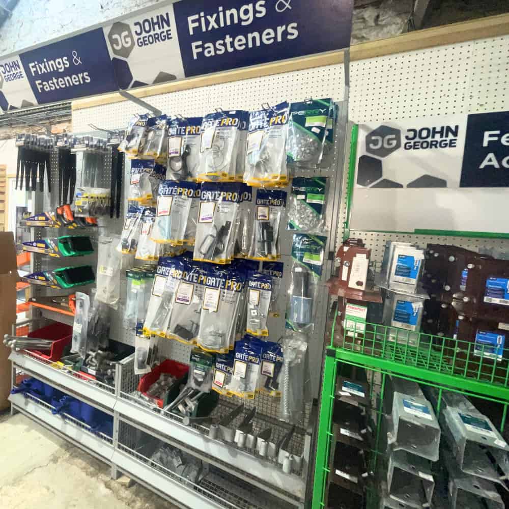 A hardware store aisle with racks displaying assorted fixings and fasteners in plastic packages, labeled under a Fixings & Fasteners sign from the brand John George. Bins of loose hardware are seen below.