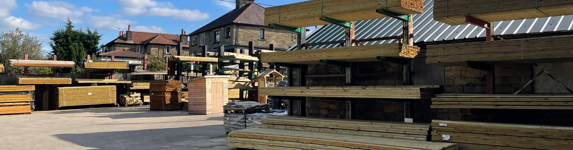Stacks of wooden planks and timber beams arranged on shelves in an outdoor lumber yard, with a few buildings and trees visible in the background under a partly cloudy sky.