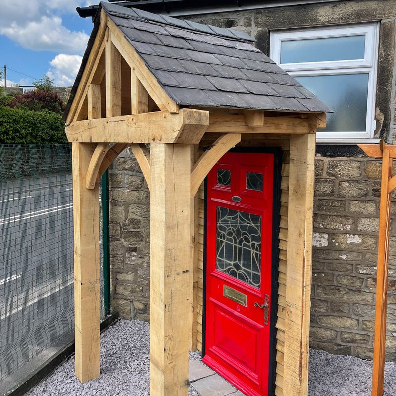Load image into Gallery viewer, The Bingley Fencing Entrance Porch in Oak features unfinished timber beams with a sloped shingled roof, standing on gravel beside a window and fence, and adds charm to any stone building with its bright red front door.

